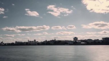 Beyaz bulutların şehrin üzerinde aktığı yaz manzarası ve geniş bir nehir, zaman ayarlı etki. Video kaydı. Kasaba, rüzgarlı bir günde nehir kıyısında yer alır..