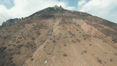 Arka planda bulutlar olan mavi gökyüzü üzerinde çalılar olan bir yaz tepesi manzarası. Vuruldu. Kamera dik dağ yamacından yukarıya, tepesine doğru hareket ediyor..
