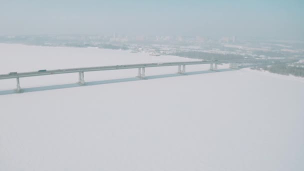 Αεροφωτογραφία μιας μεγάλης γέφυρας πάνω από χιόνι και πάγο σκεπασμένο ποτάμι μεταξύ δύο τμημάτων της πόλης. Κλιπ. Χειμερινό τοπίο με ένα ευρύ λευκό ποτάμι και μια γέφυρα με την οδήγηση αυτοκινήτων. — Αρχείο Βίντεο