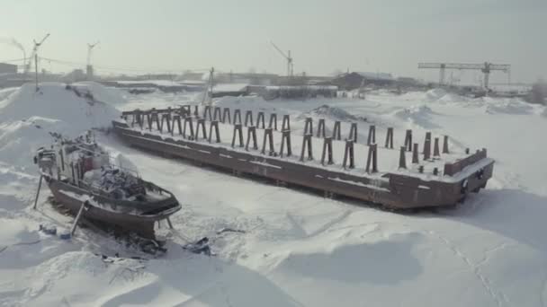 Вид с воздуха на старый пирс и пришвартованный ржавый корабль на заснеженном берегу. Клип. Зимний пейзаж заброшенного пирса и судна на фоне кранов и строительной площадки. — стоковое видео