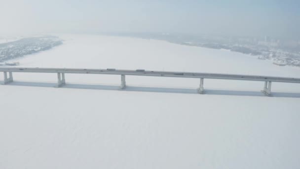 Luchtfoto van een lange brug boven sneeuw en ijs overdekte rivier tussen twee delen van een stad. Een knip. Winterlandschap met een brede witte rivier en een brug met rijdende auto 's. — Stockvideo