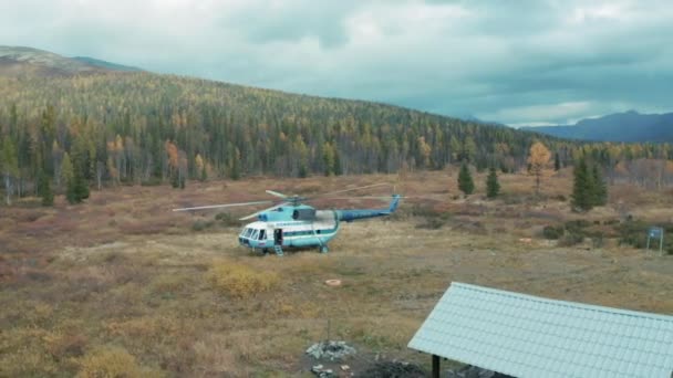 Aerial view of a helicopter with rotating blades landed on a field near countryside wooden house. Clip. Natural background with a helicopter on a meadow surrounded by forested hills and cloudy sky. — Stock Video