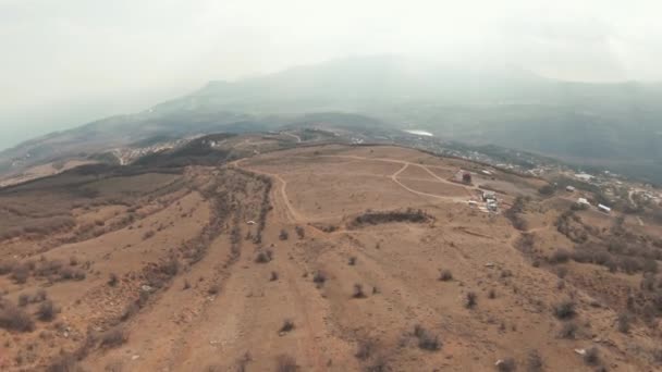 Malebný pohled na hornatou krajinu nad vesnicí na zamračeném pozadí oblohy. Zastřelen. Kamera dron pohybující se dolů podél suché půdy svahu se zeleným údolím daleko v dálce. — Stock video