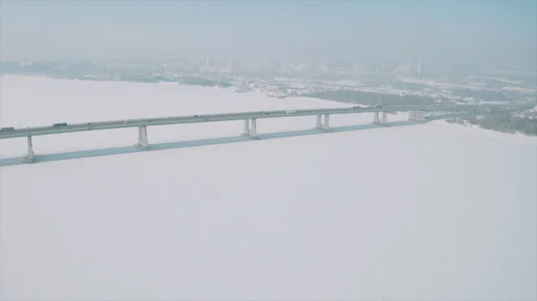 Veduta aerea di un lungo ponte sopra neve e ghiaccio coperto fiume tra due parti di una città. Clip. Paesaggio invernale con un ampio fiume bianco e un ponte con auto guida. — Foto Stock