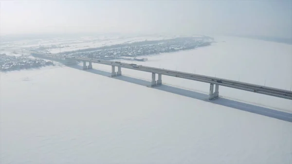 Widok z lotu ptaka długiego mostu nad śniegiem i pokrytą lodem rzeką między dwiema częściami miasta. Klip. Zimowy krajobraz z szeroką białą rzeką i most z samochodami. — Zdjęcie stockowe