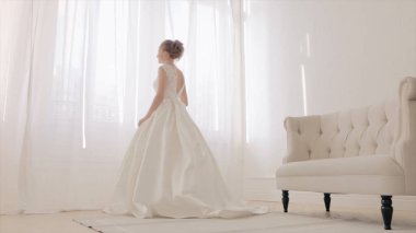 Döngülü beyaz zarif gelinlik içinde gelinin arkası. Video kaydı. Tam boy. Düğün için. Kapalı bir yerde, çiçekli güzel bir gelin. Büyük bir pencereye karşı. Uzunluk arka planda.