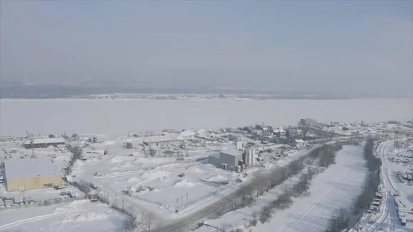 Een buitenwijk, dorp of industriële wijk van een winterstad. Een knip. Luchtfoto van een stad gelegen aan de sneeuw en ijs overdekte brede rivier. — Stockfoto