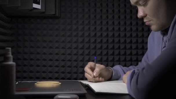 El hombre escribe en el cuaderno sobre el fondo de la pared negra. HDR. El joven escribe música en un cuaderno. El hombre escribe música o partituras en un cuaderno de estudio de grabación — Vídeo de stock