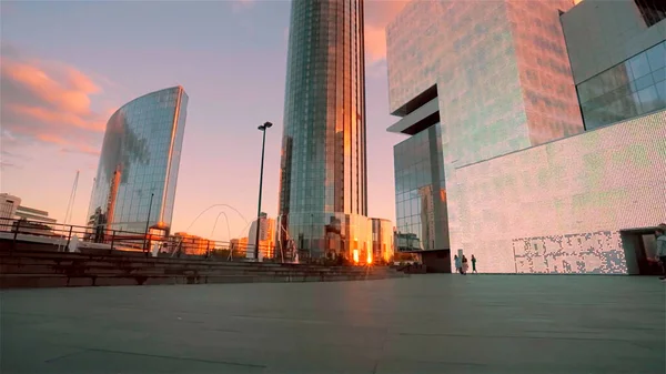 Una pequeña plaza frente a un moderno centro de negocios de fachada de vidrio con una puesta de sol dorada reflejada en las ventanas. Vídeo. Concepto de arquitectura moderna. — Foto de Stock