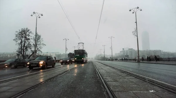 City street with driving cars and trams in late autumn season with heavy fog and falling snow. Video. Concept of traffic and big city life.