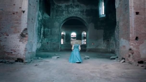 Wnętrze opuszczonego i zniszczonego kościoła z kobietą wchodzącą do środka. Nagranie. Widok na tył kobiety w tradycyjnej niebieskiej, długiej, staromodnej sukience spacerującej wewnątrz starego kościoła. — Wideo stockowe