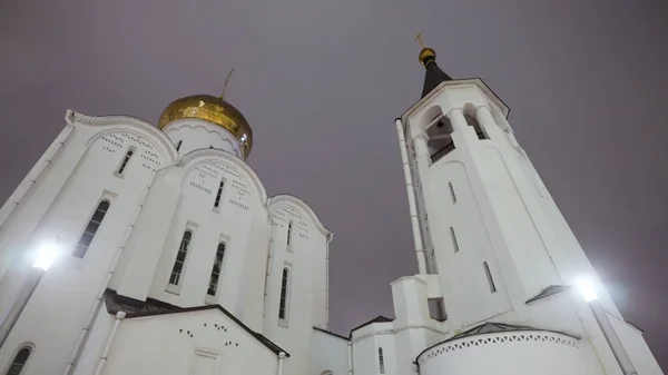 Uitzicht op een grote witte kerk met gouden koepels en een klokkentoren. Actie. Orthodoxe kerk op donkere nacht hemel achtergrond onder straatlantaarns. — Stockfoto