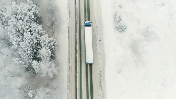 Aerial view of a logging truck driving on an empty road leading through snowy spruce forest. Scene. Concept of transportation, a truck moving along snow covered winter nature. — Stock Photo, Image