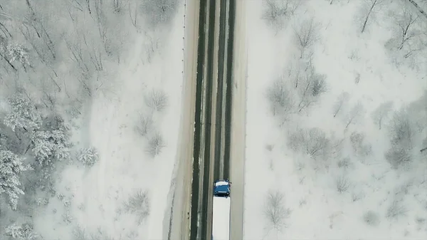 Vista aérea do tráfego na estrada que passa pela floresta de inverno em tempo severo. Cena. Vista superior de um caminhão de carga que se move em uma estrada reta cercada por madeiras cobertas de neve. — Fotografia de Stock