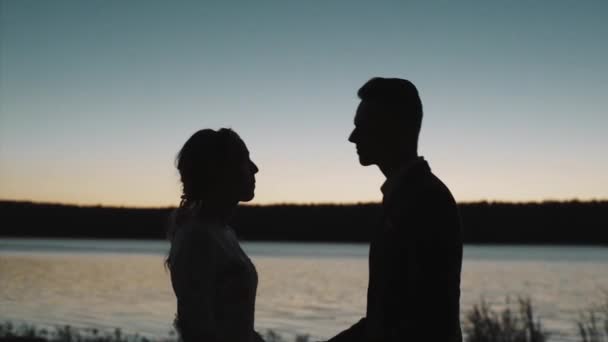 川の前の日没時に恋にカップルのシルエット。ビデオだ。ロマンチックなデート中にキス男と女のサイドビュー,愛の概念. — ストック動画