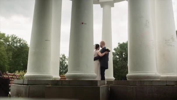 Ευτυχισμένοι νεόνυμφοι χορεύουν περιτριγυρισμένοι από τις κολώνες στο πάρκο της πόλης. Βίντεο. Νύφη και γαμπρός αγκαλιάζουν και χορεύουν σε μια καλοκαιρινή μέρα στην ύπαιθρο. — Αρχείο Βίντεο