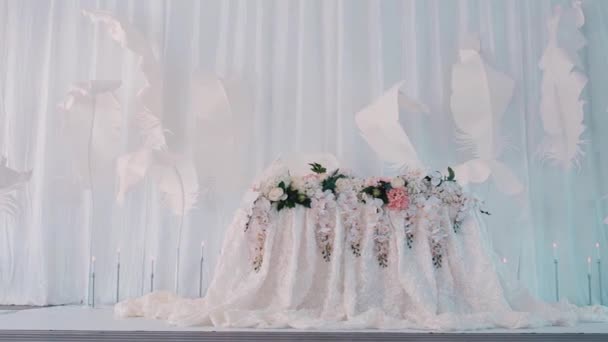 Weißes Hochzeitspräsidium mit Blumen vor weißen Wänden mit Federn. Video. Konzept des Feierns und der Liebe, ein Tisch für Frischvermählte im Saal. — Stockvideo