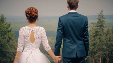 Yeşil orman vadisinin tepesindeki bir dağın tepesinde duran sevimli, genç, beyaz, yeni evli gelin ve damat. Doğal manzaranın önündeki bir çiftin arka görüntüsü..