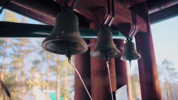 Großaufnahme von drei Glocken auf dem Hintergrund von verschwommenem blauem Himmel und Bäumen. Video. Eisenglocken hängen in einem Holzbogen. — Stockvideo