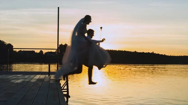 Side view of crazy and funny bride and groom jumping into the river from a pier on sunset sky background. Video. Happy husband and wife jump into the water in the summer evening. — Stock Photo, Image