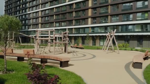 Paisaje de verano con parque infantil moderno vacío en un nuevo complejo residencial moderno. Vídeo. Parque infantil de madera con una casa de varios pisos en el fondo. — Vídeos de Stock