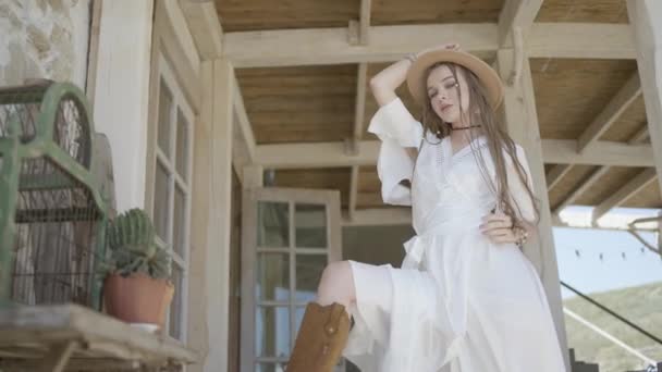 Schöne junge Frau posiert im Kleid auf der Veranda. Handeln. Sommerbild einer jungen Frau in Kleid und Cowboyhut. Junge Frau in weißem Kleid posiert vor Holzhaus — Stockvideo