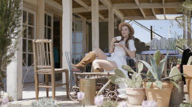 Verandada çay içen güzel genç bir kadın. Başla. Güzel elbiseli çekici kadın ahşap evde çay içiyor. Çiftlikte kovboy tarzı giyinmiş güzel bir kadın.