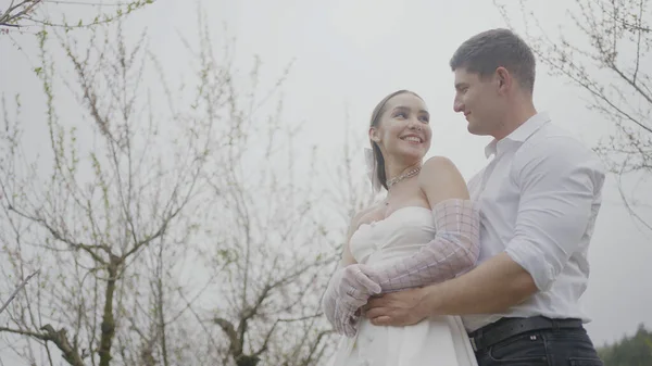 Newlyweds hug on background of sky. Action. Happy couple of newlyweds are gently embracing on background of overcast sky. Beautiful husband and wife are smiling happily in wedding dresses — Stock Photo, Image