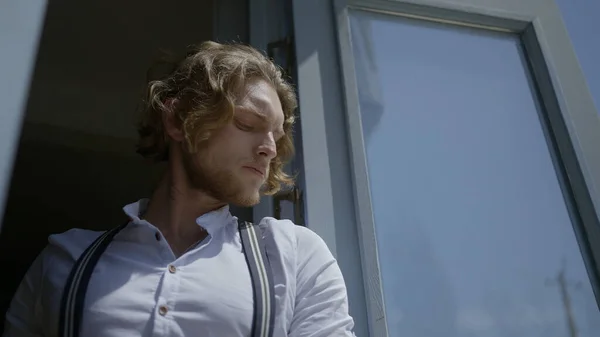 Handsome man is standing at window or door. Action. Young curly-haired man poses at entrance to house. View of stylish and attractive man standing at window or door — Stock Photo, Image