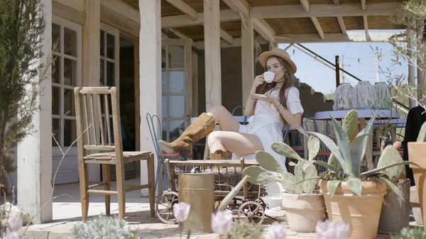 Belle jeune femme buvant du thé sur la véranda. L'action. Attrayant femme en belle robe boit du thé à la maison en bois. Belle femme en robe et style cow-boy sur ranch — Photo