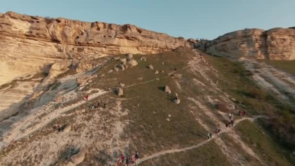 Luchtfoto van toeristische groep mensen lopen rond witte gigantische klif op blauwe heldere hemel achtergrond. Neergeschoten. Het verkennen van prachtige natuurlijke formaties met groene helling. — Stockvideo