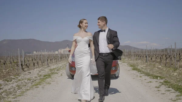 Los recién casados están caminando maravillosamente en el campo. Acción. Los recién casados con estilo se están abrazando en la carretera. Los recién casados con trajes elegantes están caminando épicamente a lo largo de la carretera de campo — Foto de Stock