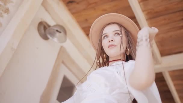 Modelo de moda feminina em vestido branco e chapéu de cowboy em pé perto da casa. Acção. Jovem posando em um dia quente de verão, vista inferior. — Vídeo de Stock