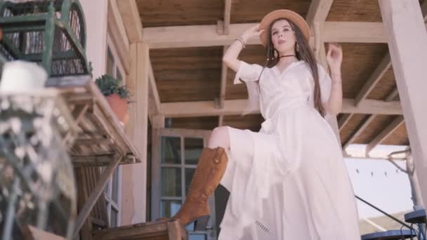 Venkovská dívka na zadní verandě, bílé šaty a kovbojský klobouk. Akce. Dolní pohled na pózující modelku s nohou v hnědé kovbojské botě na dřevěném stole. — Stock video