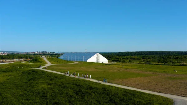 Aerial view of a green meadow and a white industrial greenhouse on blue sky background. Video. People walking on a narrow path in the countryside ecological area.