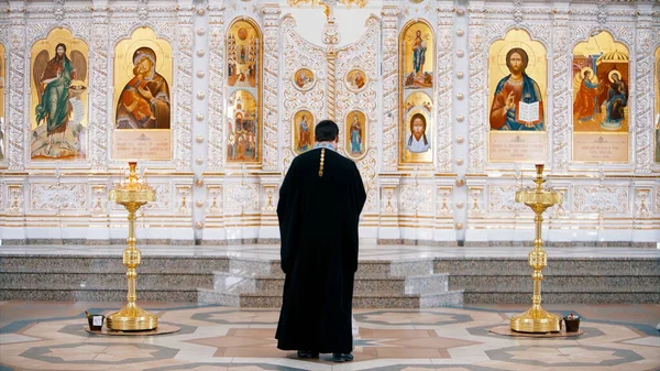 Az ikonosztázis egy ortodox templomban. Videó. Hátsó nézet egy pap áll az ikonok előtt a szentek arcával, a vallás fogalmával, belső részletek a templomban. — Stock Fotó