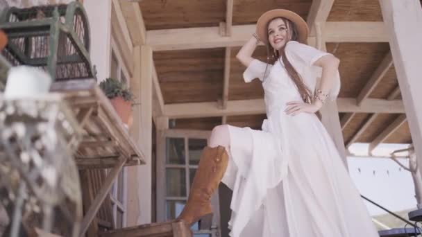 Rapariga do campo no alpendre, vestido branco e chapéu de cowboy. Acção. Visão inferior de um modelo feminino posando com a perna na bota de cowboy marrom em uma mesa de madeira. — Vídeo de Stock