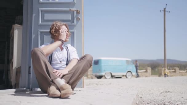 Um homem modelo relaxante e sentado em uma estrada empoeirada em um dia quente de verão. Acção. Bonito homem na frente da porta azul, céu e pequeno ônibus para viajar. — Vídeo de Stock
