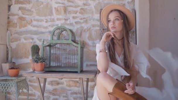 Hermosa joven modelo femenina de estilo occidental sentada frente a la pared de ladrillo, cactus y una jaula de pájaros vacía. Acción. Una mujer con muchas trenzas con botas de vaquero, un sombrero y un vestido blanco. — Vídeos de Stock