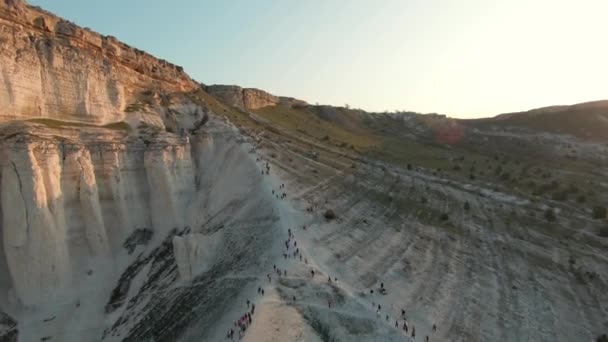 Luchtfoto van toeristische groep mensen lopen rond witte gigantische klif op blauwe heldere hemel achtergrond. Neergeschoten. Het verkennen van prachtige natuurlijke formaties met groene helling. — Stockvideo