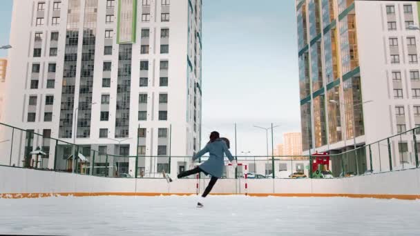 Patineuse professionnelle pratiquant le patinage artistique sur une patinoire extérieure. Vidéo. Femme danse sur glace entourée de maisons de grande hauteur résidentielles. — Video