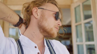 Gözlüklü acımasız genç adam. Başla. Sakallı bir adam saçlarını eliyle düzeltir. Kulübenin arka planında siyah gözlüklü seksi adam.