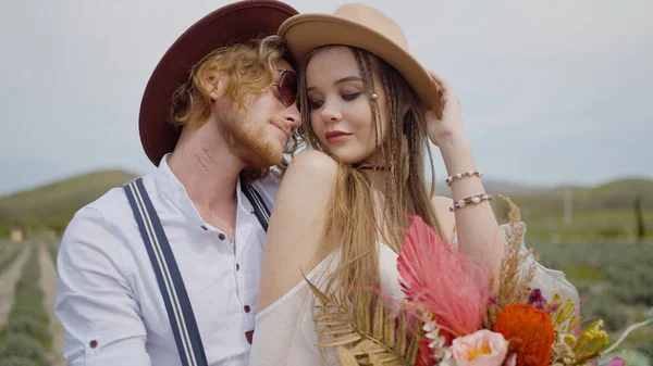 Casal atraente abraça com buquê no campo. Acção. Casal elegante de hippies ou hipsters estão abraçando no campo. Casal no amor abraços e poses no fundo do belo campo — Fotografia de Stock