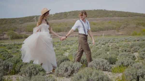 Bella coppia di sposi appena sposati cammina sul campo. Azione. Sposi in abiti chic ed eleganti stanno camminando intorno al campo. Sposo attraente conduce sposa a mano attraverso il campo — Foto Stock