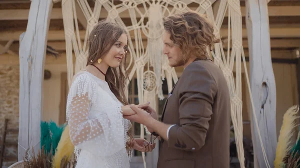 Hochzeit im Hippie-Stil. Handeln. Schönes Brautpaar tauscht Ringe unter Krone aus. Hippie-Frischvermählte verloben sich in schönen Outfits vor dem Hintergrund der geflochtenen Krone — Stockfoto