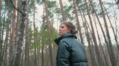 Güzel genç bir kadın yeşil ormanda yürüyor. Başla. Ceketli güzel bir kadın ormanda yürüyor ve güzelliğinin tadını çıkarıyor. Kadın ormanda yürür ve uzun ağaçlara bakar.