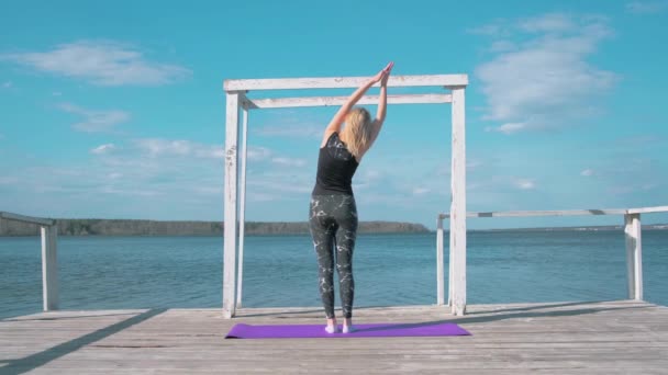 Vista trasera de la mujer en el muelle haciendo yoga. Acción. Hermosa mujer hace yoga en muelle de madera con vista al mar. La mujer hace yoga en la playa en la mañana soleada ventosa. Yoga, motivación, salud — Vídeos de Stock