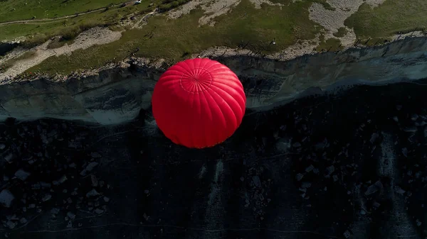 Flygfoto över en flygning av en röd varmluftsballong över klipporna på landsbygden. Skjuten. Flyger ovanför den steniga bergssluttningen, gröna ängen och en smal stig. — Stockfoto
