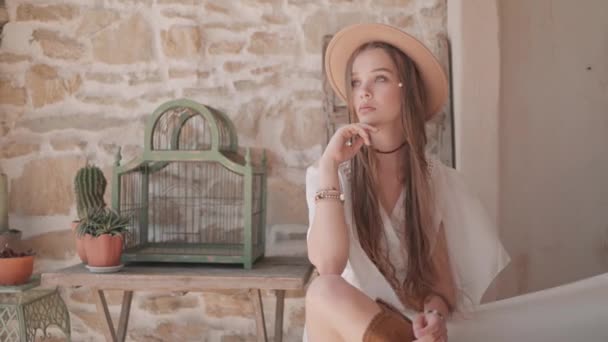 Hermosa joven modelo femenina de estilo occidental sentada frente a la pared de ladrillo, cactus y una jaula de pájaros vacía. Acción. Una mujer con muchas trenzas con botas de vaquero, un sombrero y un vestido blanco. — Vídeos de Stock