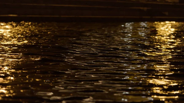 Primer plano de la superficie del agua con las luces de la ciudad nocturna. Imágenes de archivo. Rippled amplia rive y la calle oscura de la ciudad en una noche de verano con gente caminando. —  Fotos de Stock